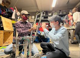 Builders (Aditya and Ryan) work on large (18 inches by 18 inches) metal robot. One member (Aditya) on the left, holds up linears on the front of the bot up for the other member (Ryan) who screws an outtake box