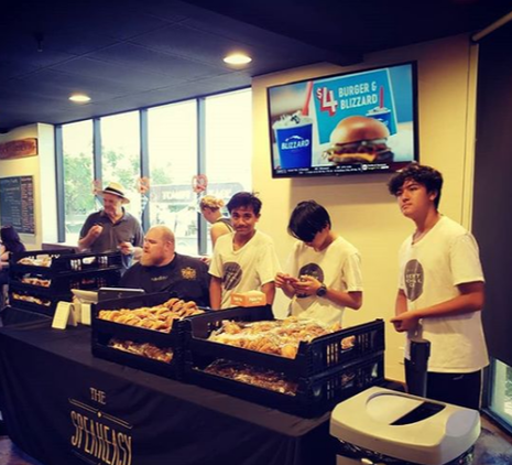 Three members stand in front of a black table with baked goods on it which they are helping to sell. Behind them is a TV with a $4 Burger and Blizzard ad