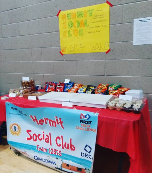 A red table in front of a grey brick wall has chips and baked goods on it for a bake sale we did at the Makerspace Festival. On the red table, hanging on the front, is a Hermit Social Club Banner